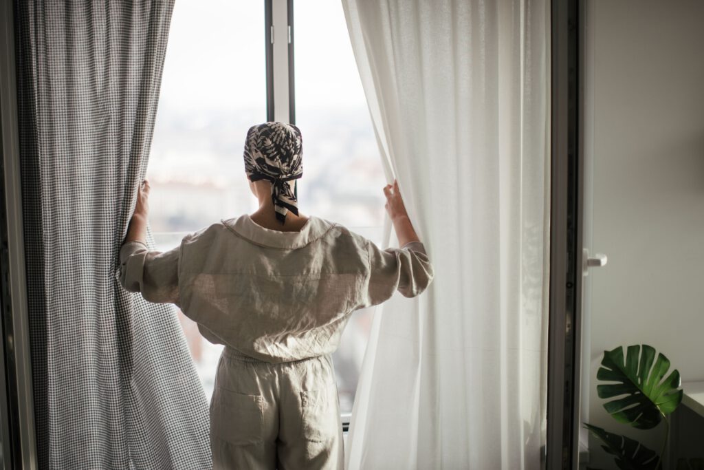 Rear view of young unhappy woman with cancer standing in front of a window.