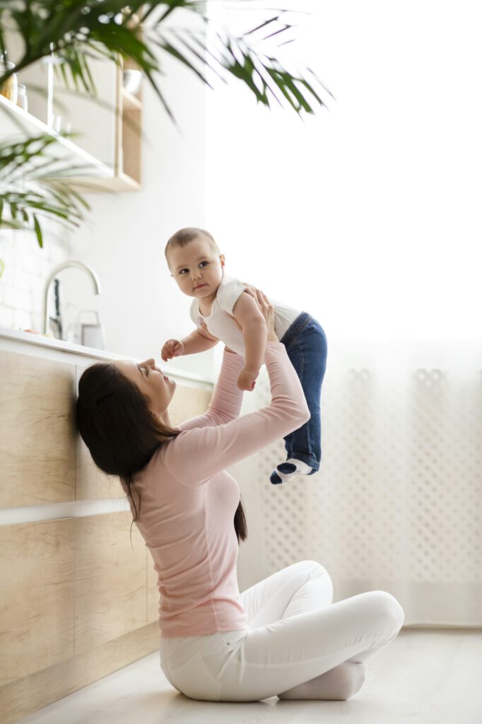 Young mom lifting her baby son up in air, sitting on kitchen floor